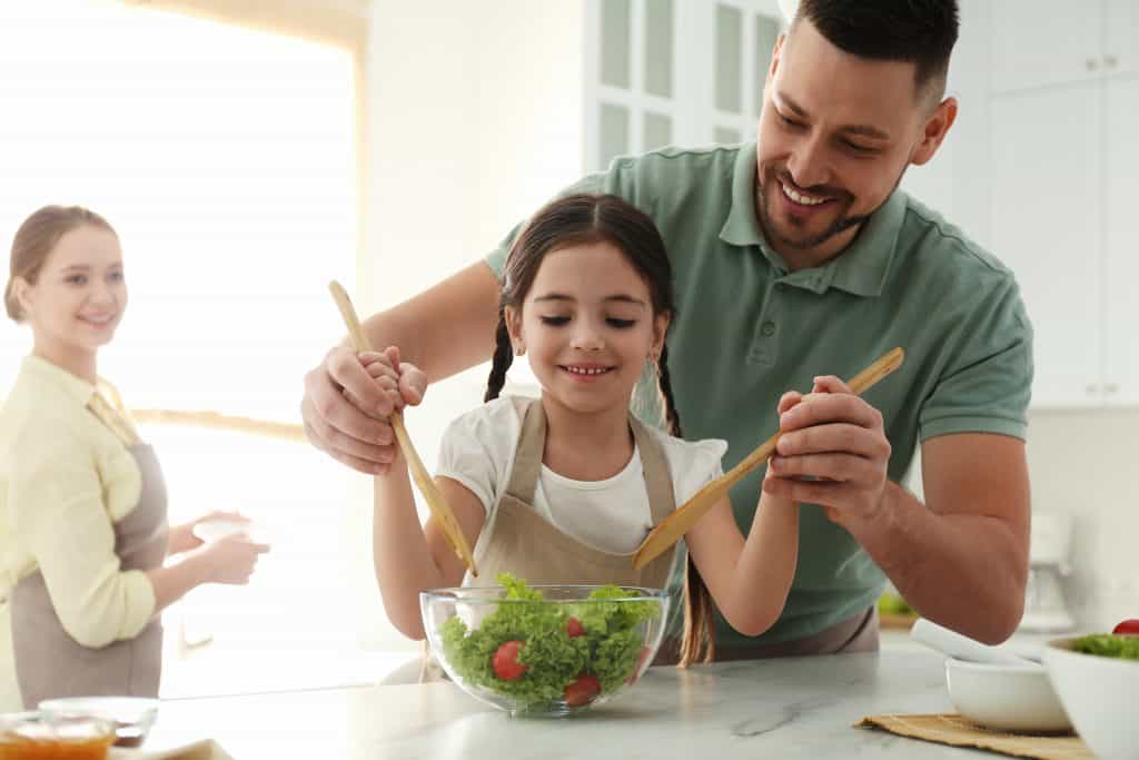 Father and daughter enjoying mixing a bowl of salad with the mother watching at the back.